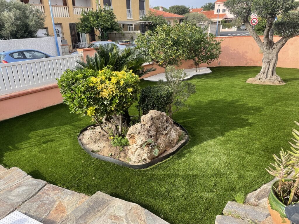 Gazon synthétique et olivier : aménagement de jardin moderne. Jardin méditerranéen avec pelouse synthétique verte éclatante, grand olivier et massif central rocheux. Mur saumon en arrière-plan et panneau 30.