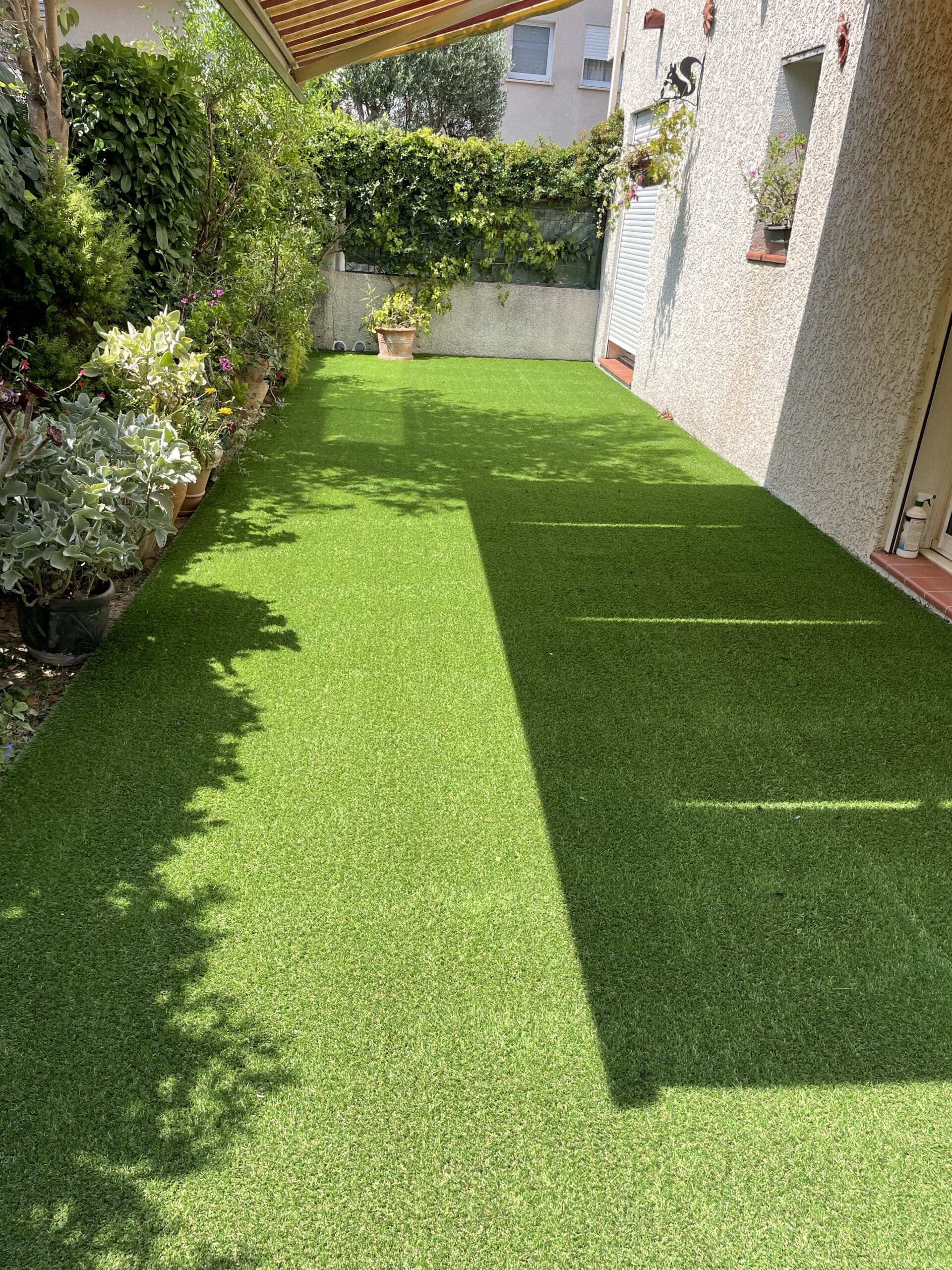 Gazon synthétique vert vif dans un jardin urbain ensoleillé. Longue bordure de plantes en pot et mur texturé. Forte ombre diagonale.