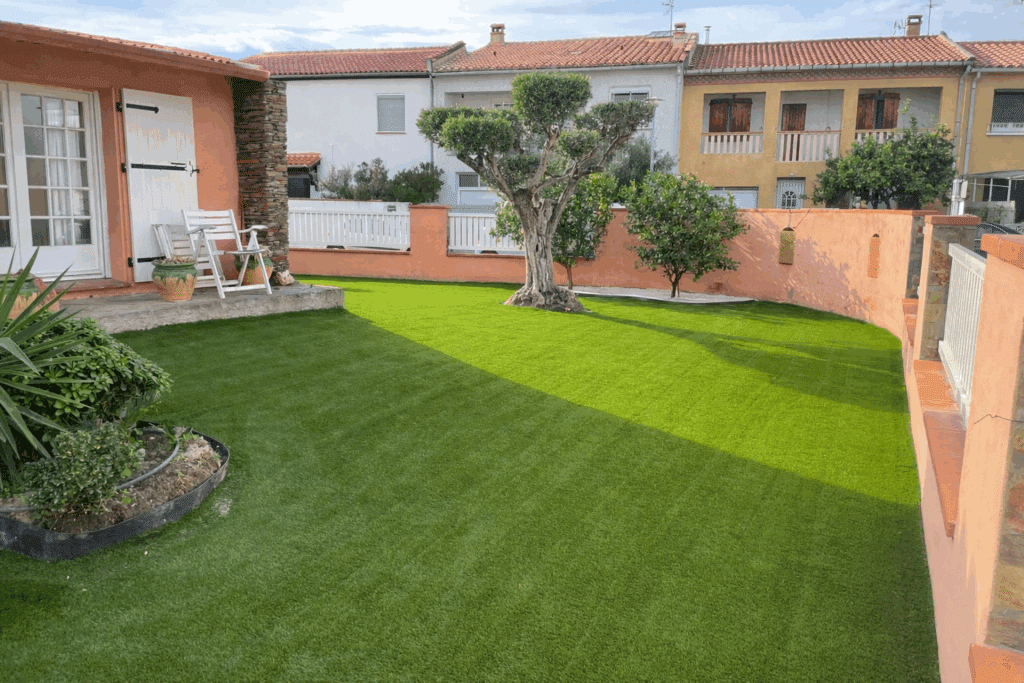 Gazon synthétique pour jardin méditerranéen moderne Jardin méditerranéen ensoleillé. Grand olivier sur gazon synthétique vert. Patio avec chaises blanches, murs saumon et maisons voisines.