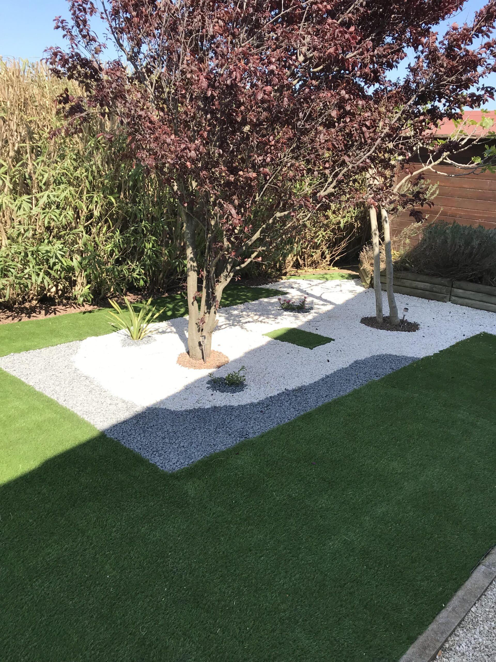 Aménagement paysager contemporain style zen. Graviers décoratifs blancs et gris délimitent l'allée entre le gazon synthétique et les arbres.