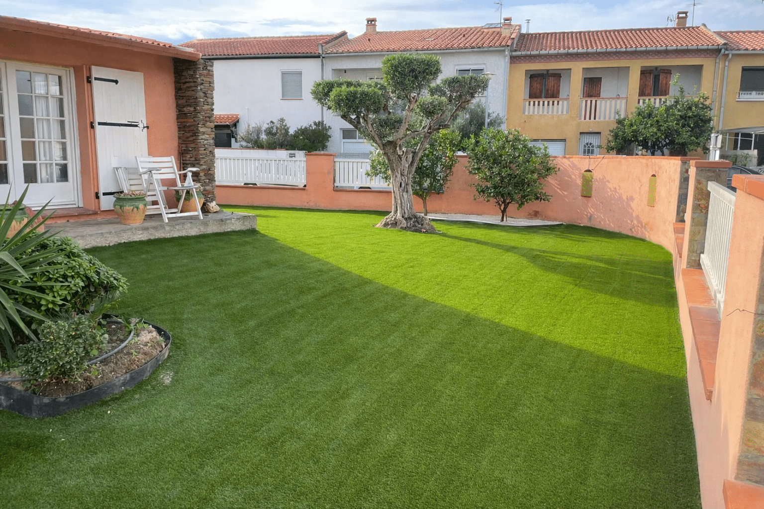 Jardin ensoleillé aménagé avec gazon synthétique vert vif. Mur terracotta, vieil olivier taillé et petite terrasse avec chaises.