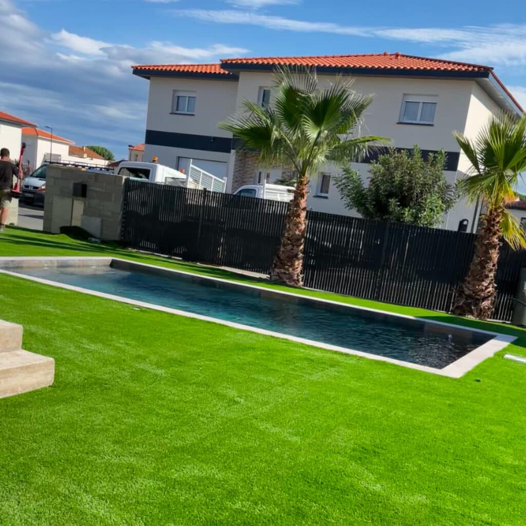 Gazon synthétique et piscine : Aménagement extérieur moderne. Couloir de nage moderne dans un jardin résidentiel. Pelouse artificielle verte, palmiers et maison blanche avec toit en tuiles rouges.