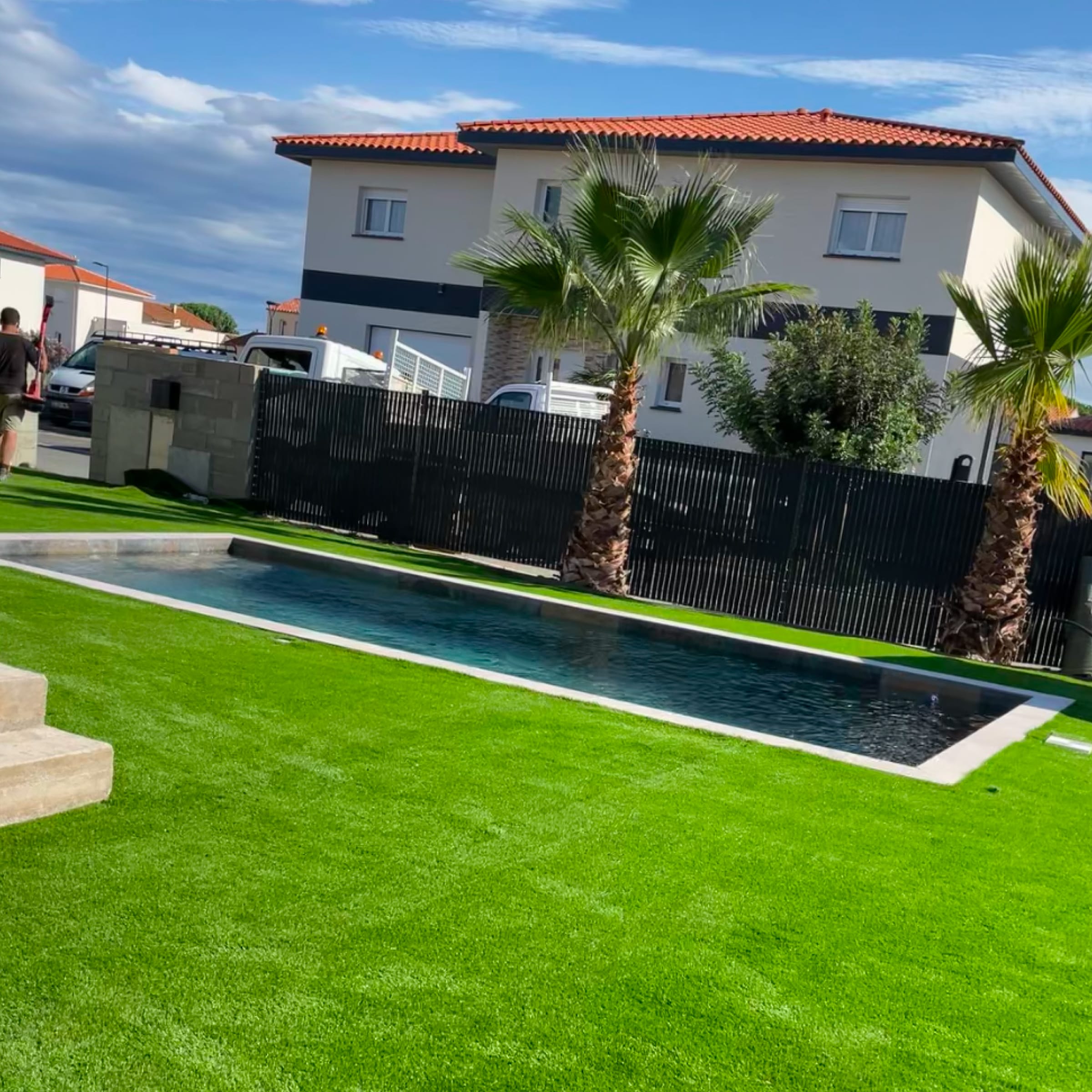 Couloir de nage moderne dans un jardin résidentiel. Pelouse artificielle verte, palmiers et maison blanche avec toit en tuiles rouges.