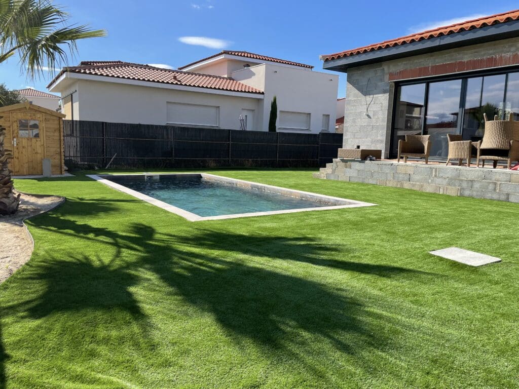 Piscine et gazon synthétique : Aménagement de jardin moderne Aménagement extérieur moderne: piscine rectangulaire, grande surface de gazon synthétique, terrasse en pierre, palmier et maisons.